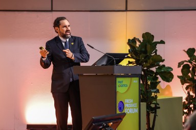 A man presents on stage at the Fresh Produce Forum during Fruit Logistica, gesturing with a device in his hand.