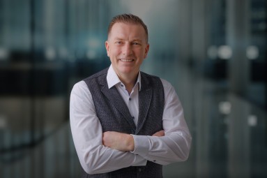 A man in a modern office, white shirt, dark waistcoat. With arms folded, he looks friendly at the camera.