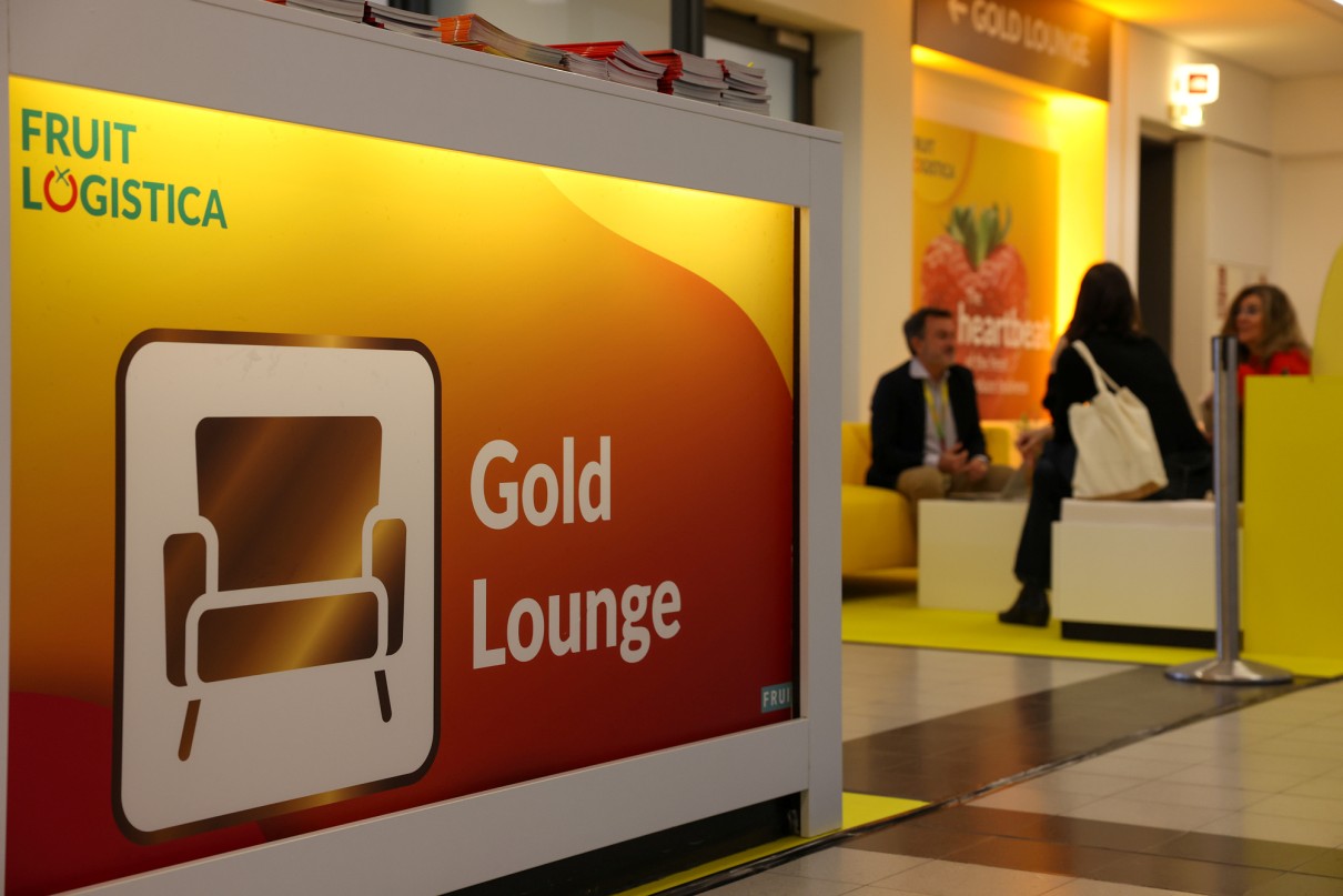 A sign with the inscription 'Gold Lounge' and a stylised armchair symbol stands in the foreground. In the background, several people sit on yellow sofas in a lounge.