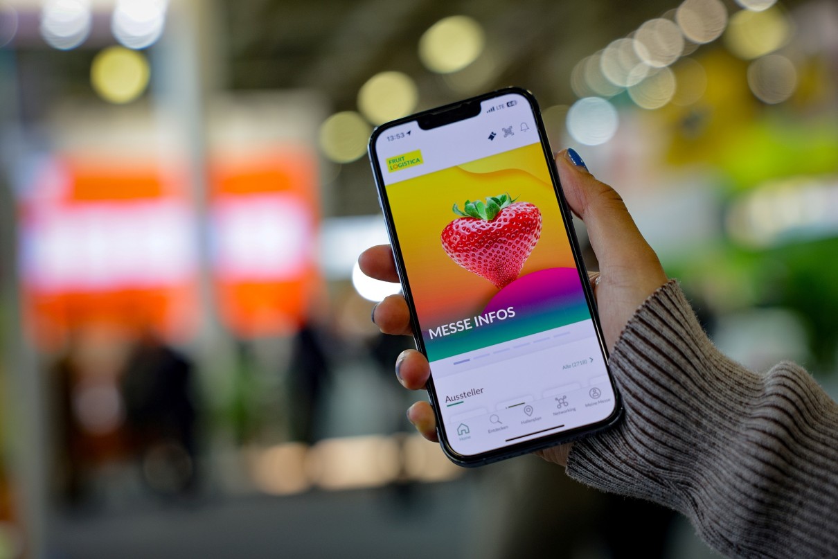 A hand is holding a smartphone. The FRUIT LOGISTICA app is open on the screen. An exhibition hall is visible in the background. 