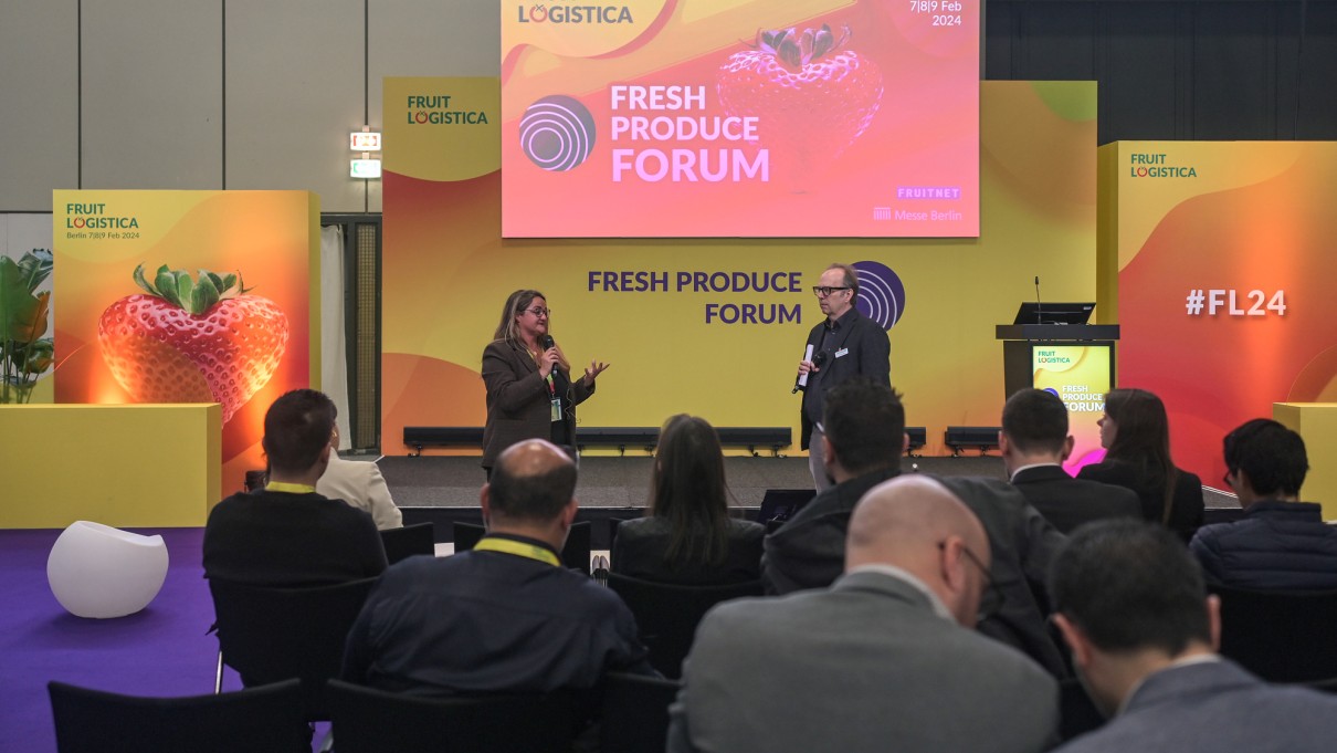 Zwei Personen halten Vorträge auf einer Bühne beim Fresh Produce Forum der FRUIT LOGISTICA 2024. Im Hintergrund gelb-orangefarbene Gestaltung mit dem FRUIT LOGISTICA-Logo und einem Erdbeer-Motiv.