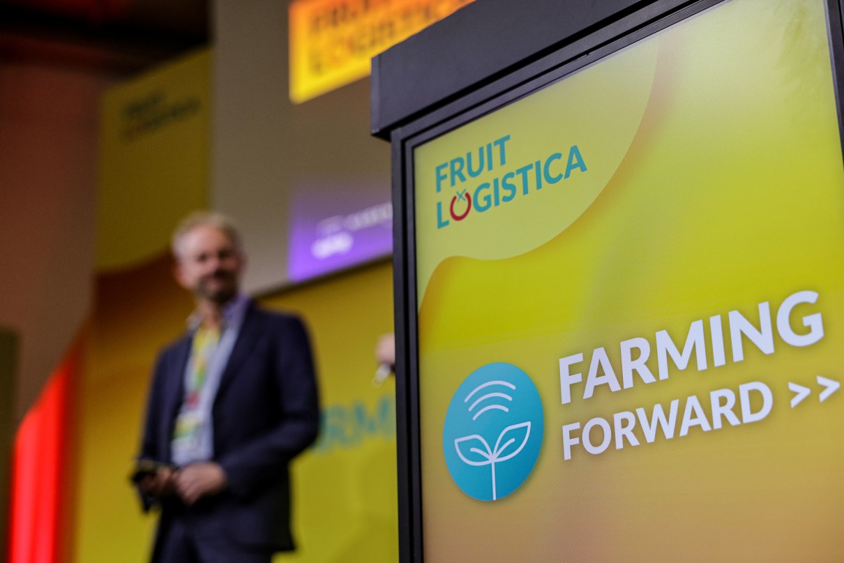 Close-up of a screen displaying ’Farming Forward’ and the FRUIT LOGISTICA logo. A man is standing onstage in the background. 