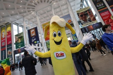 A large exhibition hall with high ceilings, full of people. In the foreground is a person in a yellow banana costume.