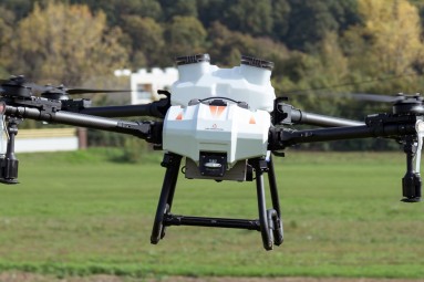 An agricultural drone with a white body and black rotor arms flying over a green field. Trees and buildings are visible in the background.