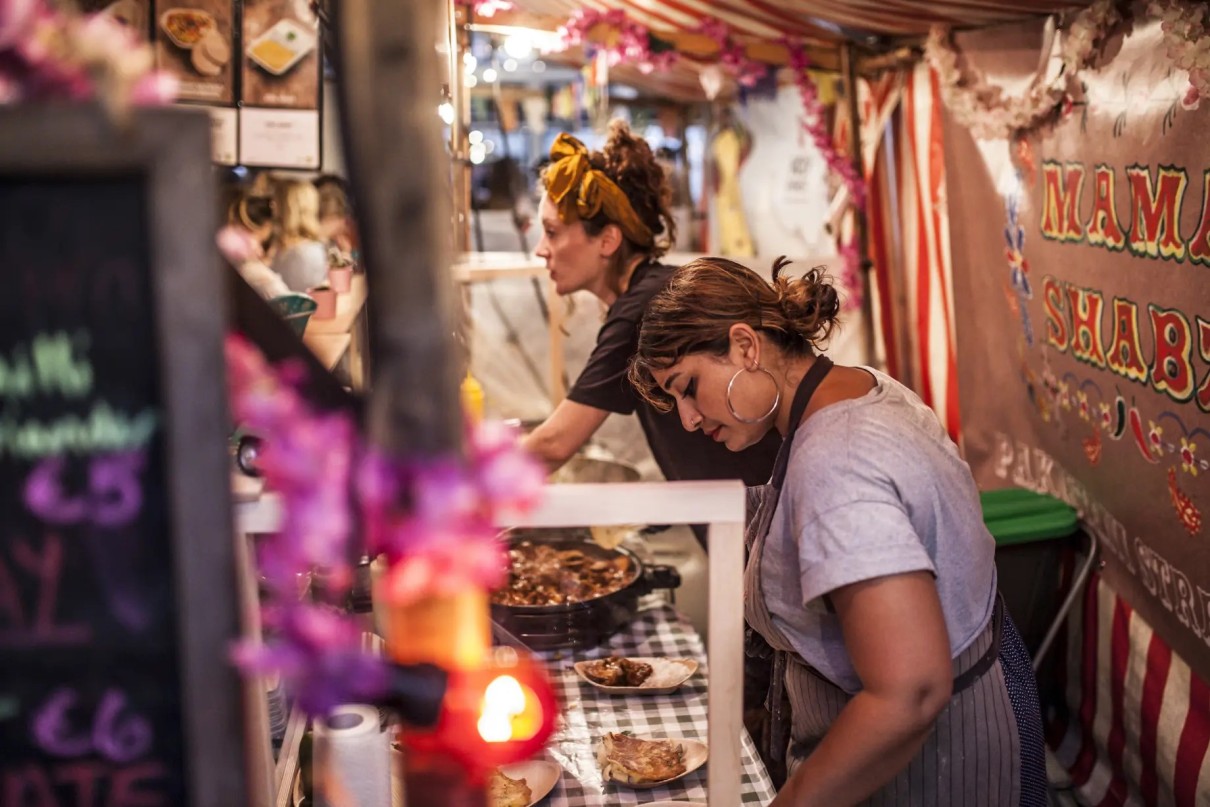 Two women are standing at a food stall in Markthalle Neun in Berlin. The scene projects a warm atmosphere with colourful decorations and a sign reading ’Mama Shabz – Pakistani Street Food’. On the cou