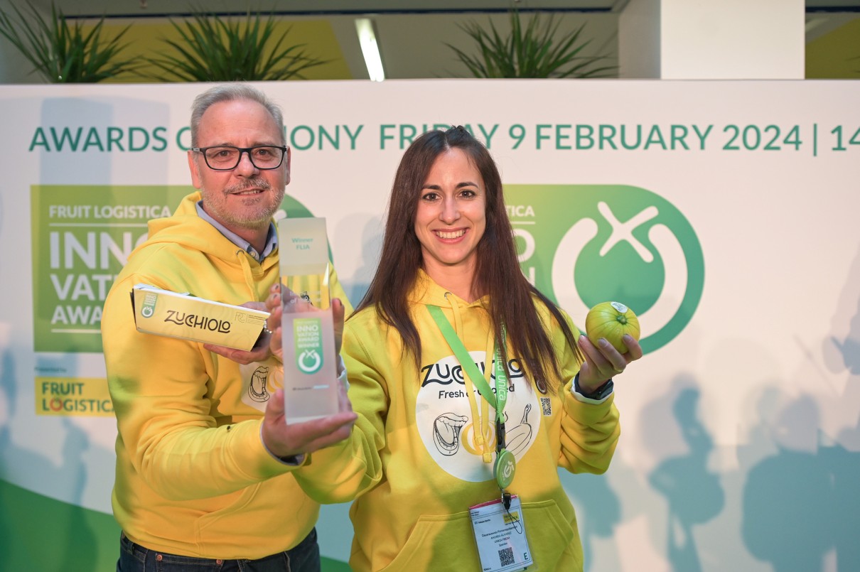 A man and a woman in yellow hoodies with the Zucchiolo logo stand smiling in front of a FRUIT LOGISTICA winners' wall. The man is holding a trophy and packaging.