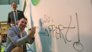 The photo shows a man writing on a large white poster with a marker pen while smiling at the camera. Standing in the background.