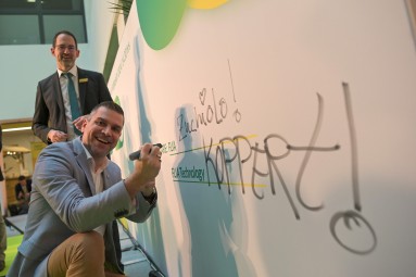 The photo shows a man writing on a large white poster with a marker pen while smiling at the camera. Standing in the background.