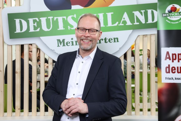 A man in a black suit stands in front of a large logo and smiles.