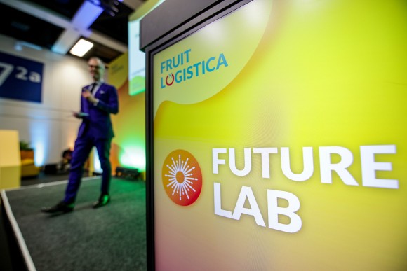 A presenter in a blue suit speaks on stage at a professional event, holding a microphone. In the foreground, a bright yellow sign reads “FRUIT LOGISTICA Future Lab”. 