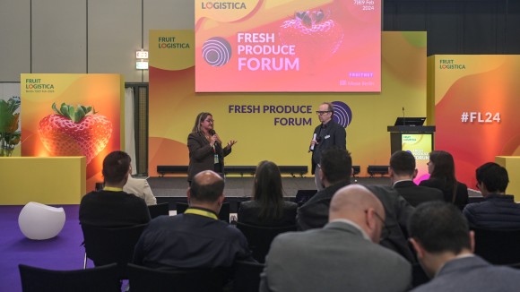 Two people are giving presentations on a Fresh Produce Forum stage at FRUIT LOGISTICA 2024. A yellow-orange design with the FRUIT LOGISTICA logo and a strawberry motif are visible in the background.